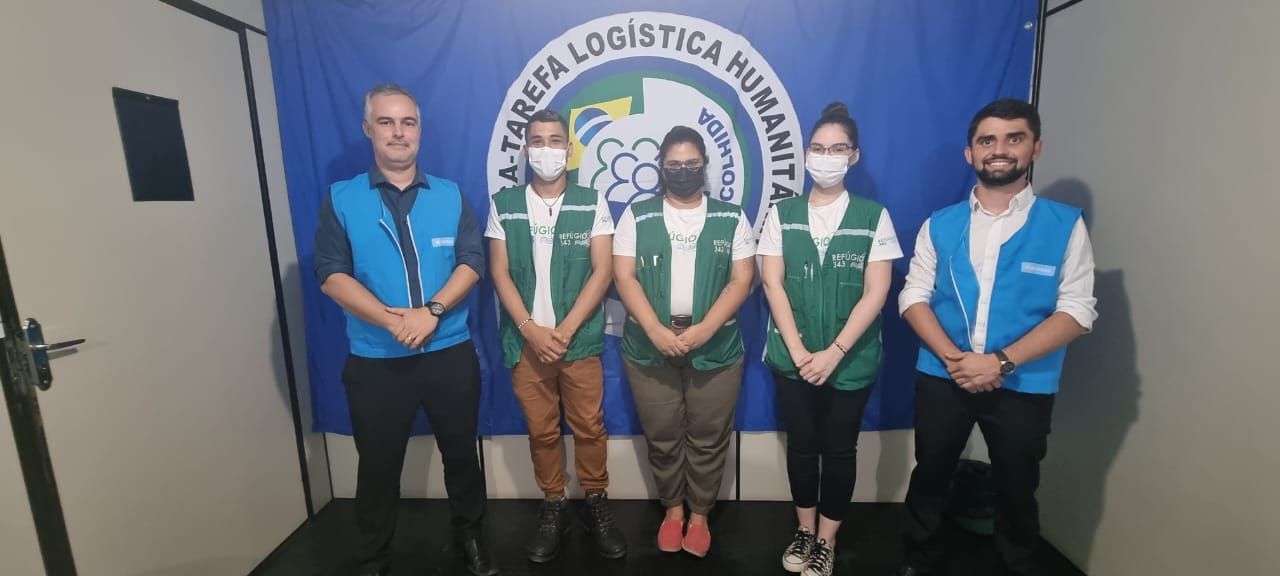 Cinco pessoas com coletes azuis posam em frente a uma faixa azul em um evento de logística.