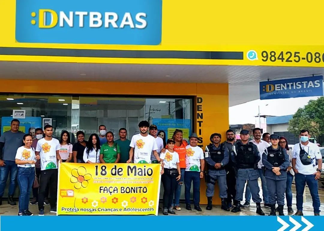 Grupo posando em frente à clínica odontológica DNTBRAS, com faixa “18 de Maio” amarelo