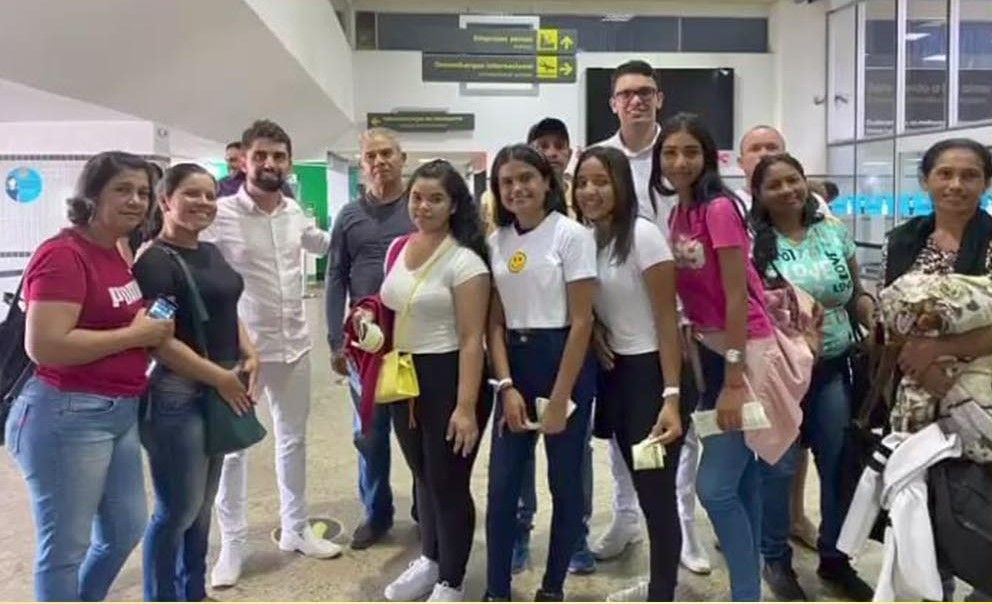 Grupo de pessoas sorrindo em um terminal de aeroporto, em pé juntas com malas e mochilas.