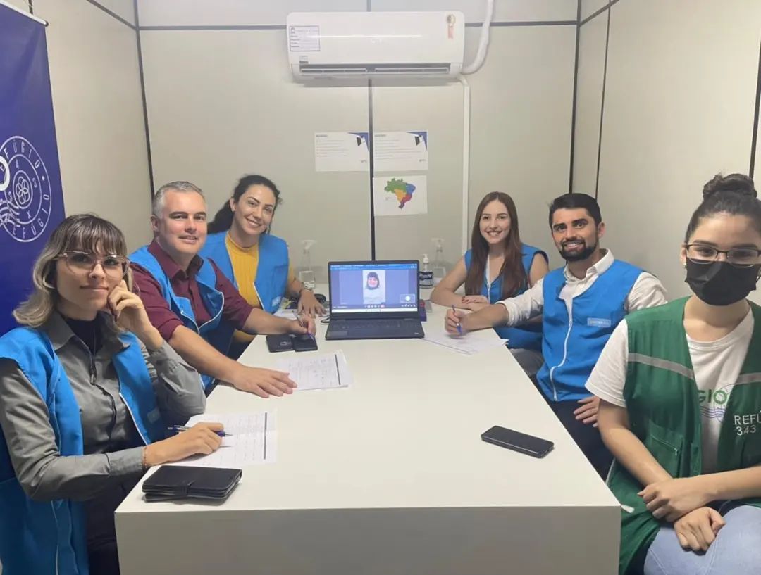 Seis pessoas com coletes azuis sentadas ao redor de uma mesa em uma sala de reuniões, olhando para um laptop.