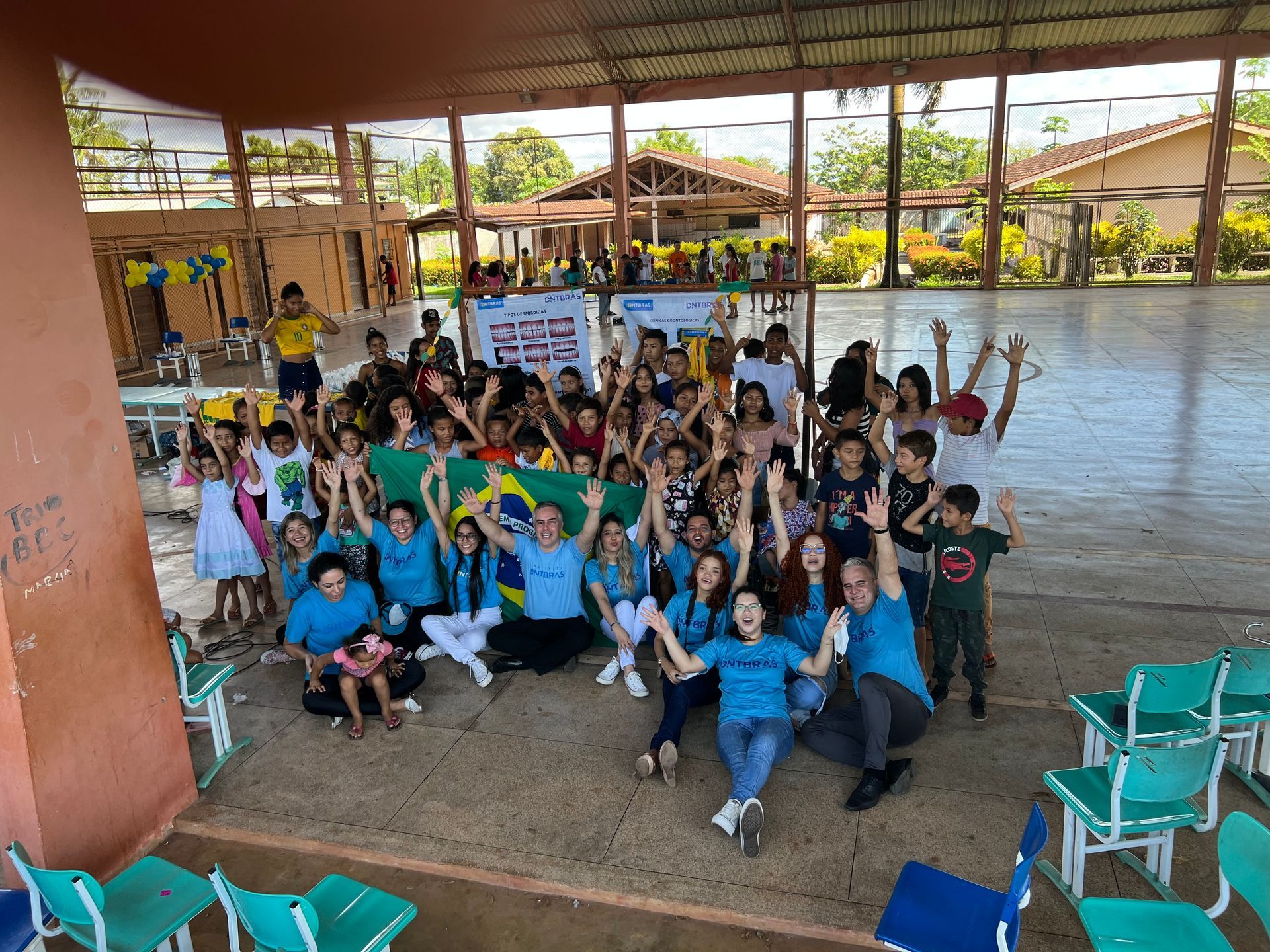 Um grande grupo posando com as mãos erguidas em um pavilhão externo coberto, com cadeiras e chão de areia.