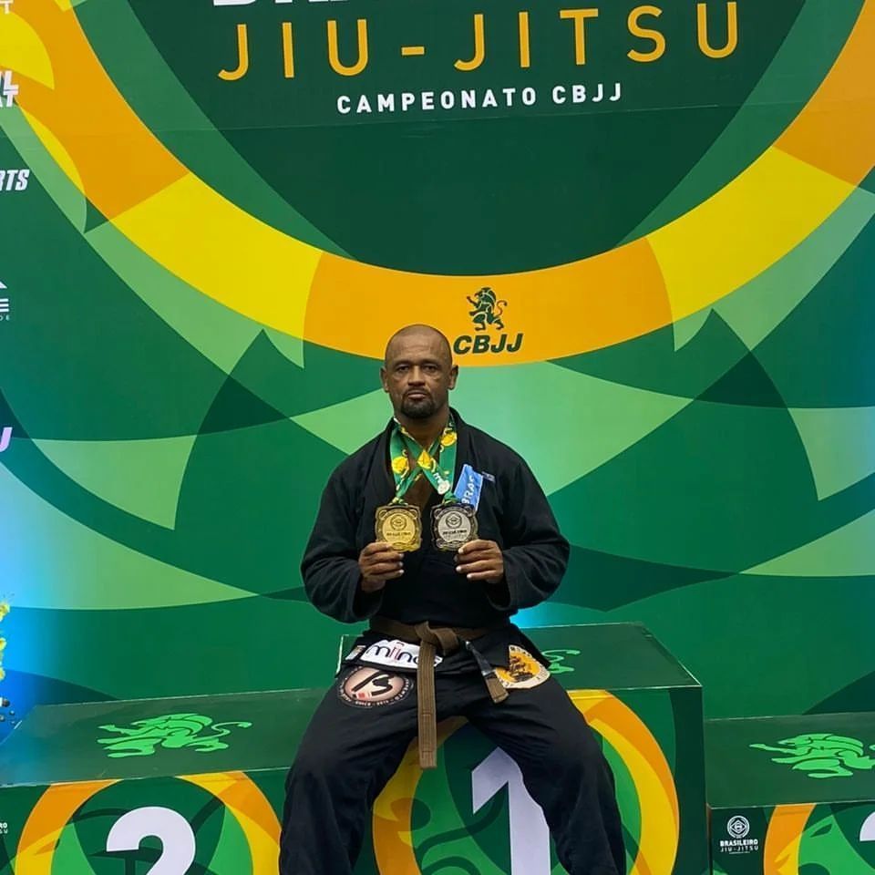 Medalhista de Jiu-Jitsu sentado no pódio segurando duas medalhas de ouro diante do cenário do campeonato CBJJ.