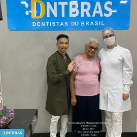 Três pessoas posando em frente a uma placa da DNTBRAS Dentistas do Brasil, uma delas usando jaleco branco e máscara.