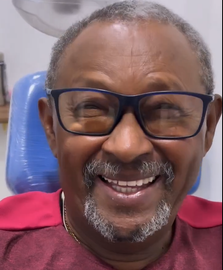 Homem sorridente, usando óculos e camisa bordô, sentado em uma cadeira azul dentro de casa.
