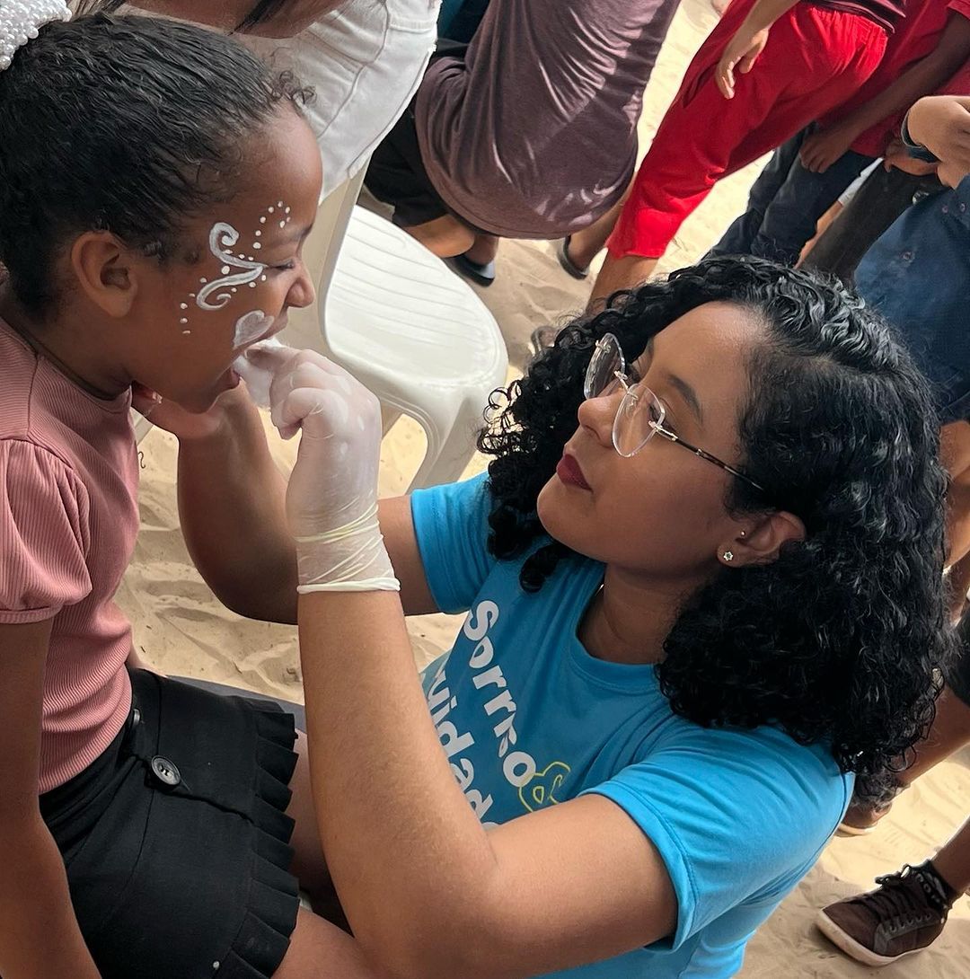 Pintura facial em andamento em um evento lotado, com uma mulher aplicando desenhos brancos no rosto de uma criança.