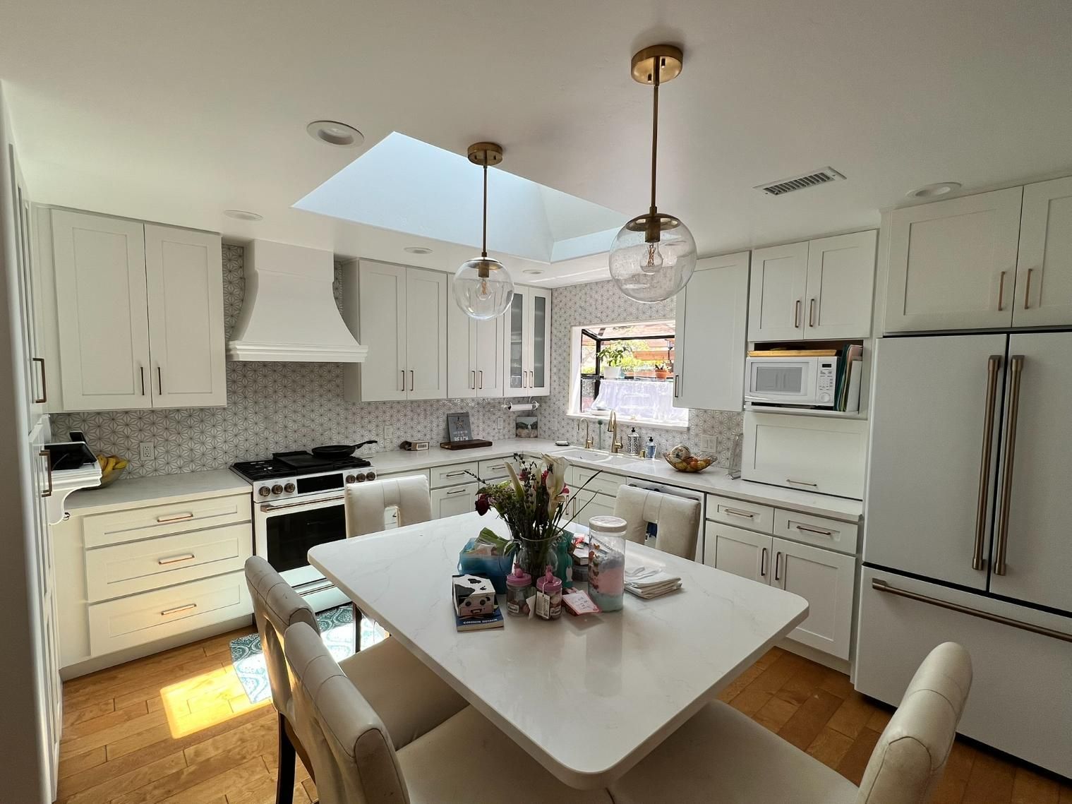Bright white kitchen with island, skylight, and stainless appliances; sunlit dining space with flowers.