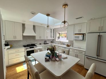Bright white kitchen with island, skylight, and stainless appliances; sunlit dining space with flowers.