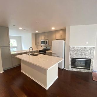 Bright modern kitchen with white island, stainless appliances, dark wood floors, and tiled fireplace wall