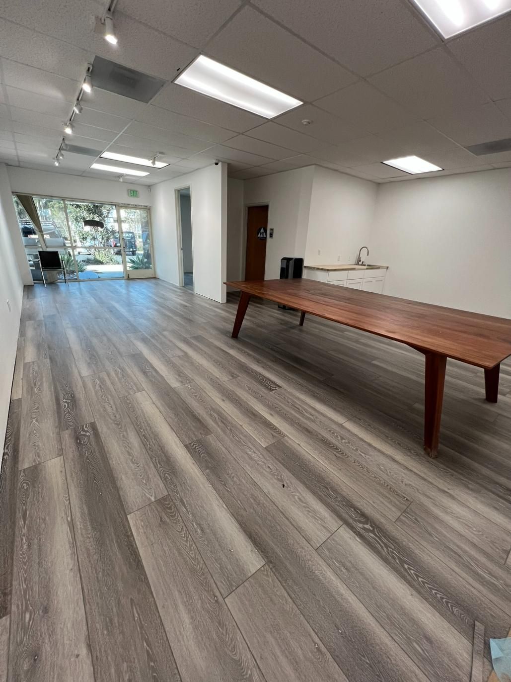 Bright empty office space with gray wood floors, white walls, ceiling lights, and a long wooden table.