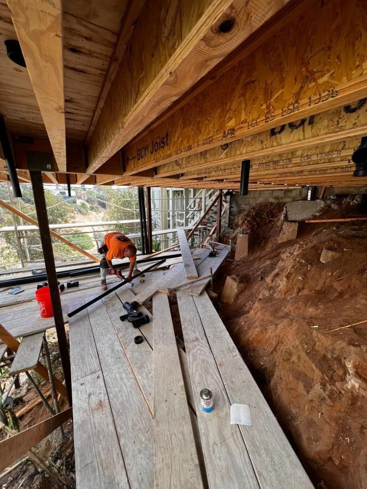 Construction worker on a narrow wooden scaffold beside a dirt slope under a deck.
