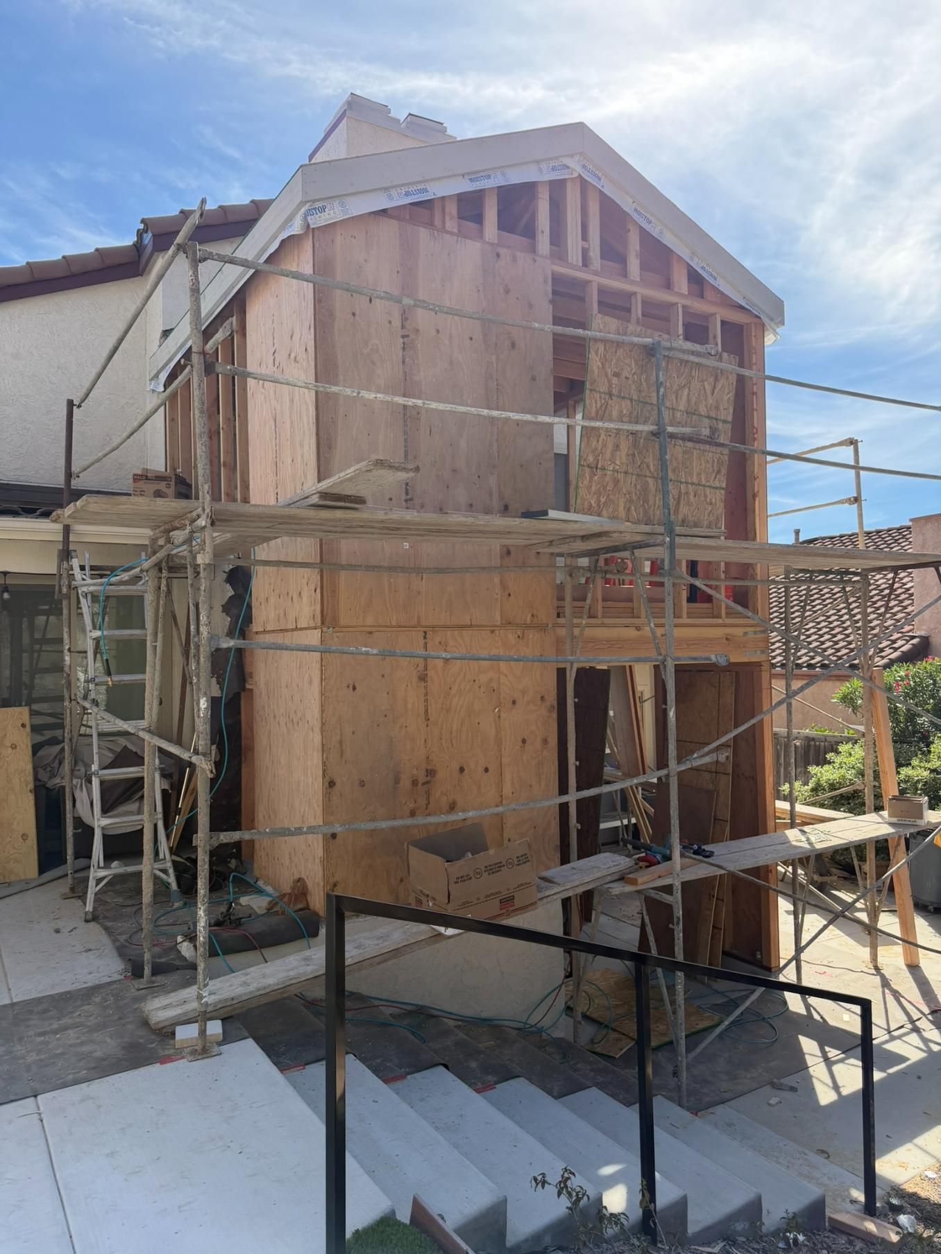 Scaffolded house under construction with plywood siding and a ladder, viewed from outside on a sunny day