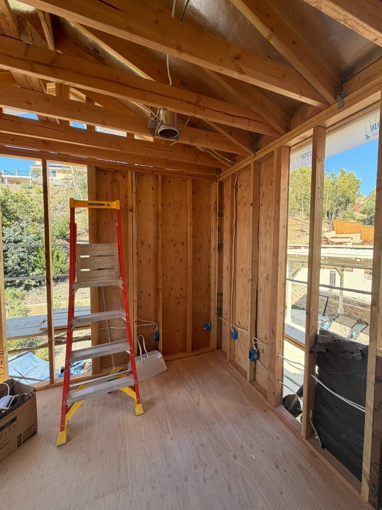 Framed wooden shed interior with ladder, open window, exposed studs, and plywood floor under construction
