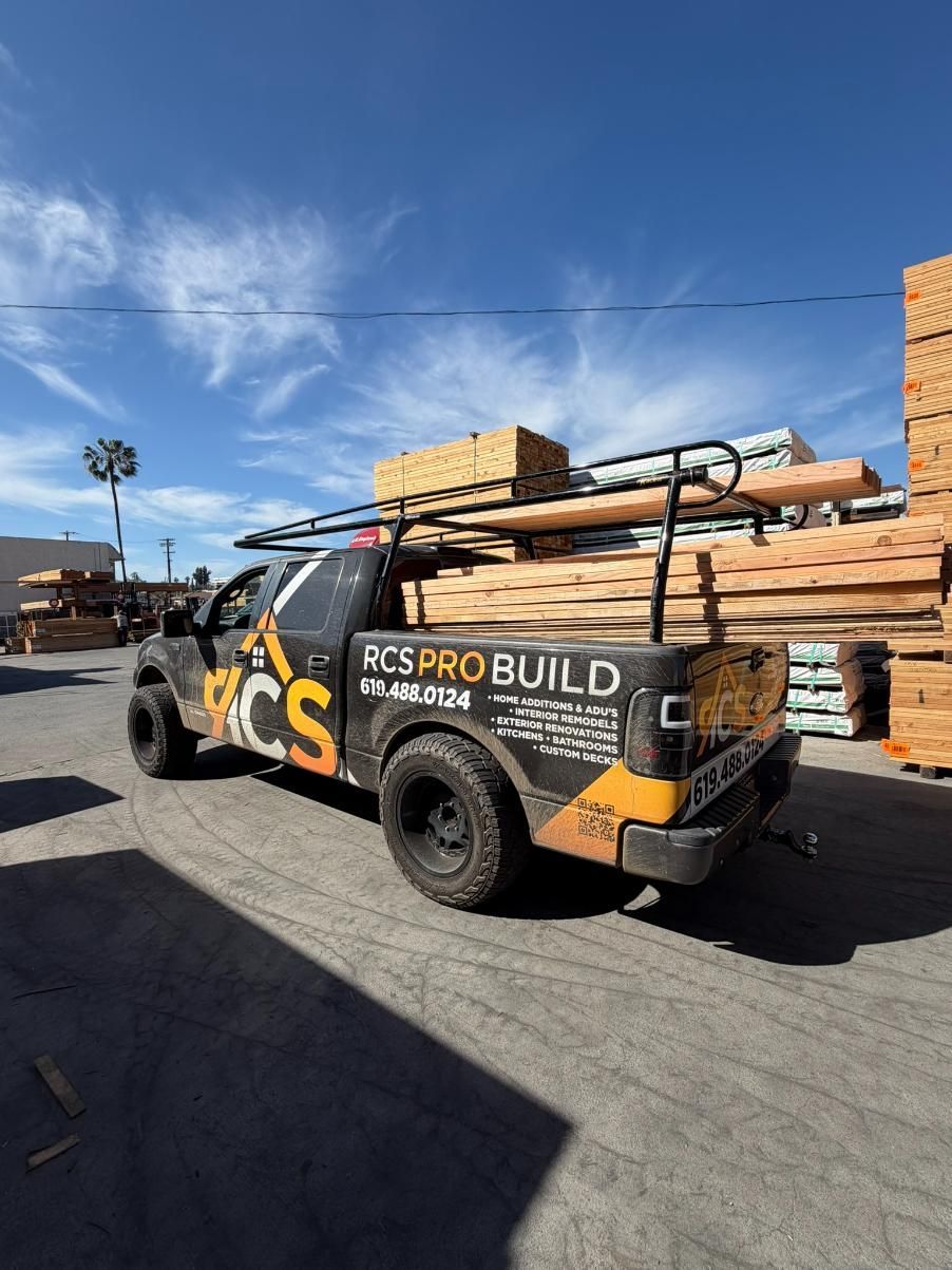 Yellow utility truck with lumber racks and “PCS Pro Build” signage parked on a sunny street under a blue sky