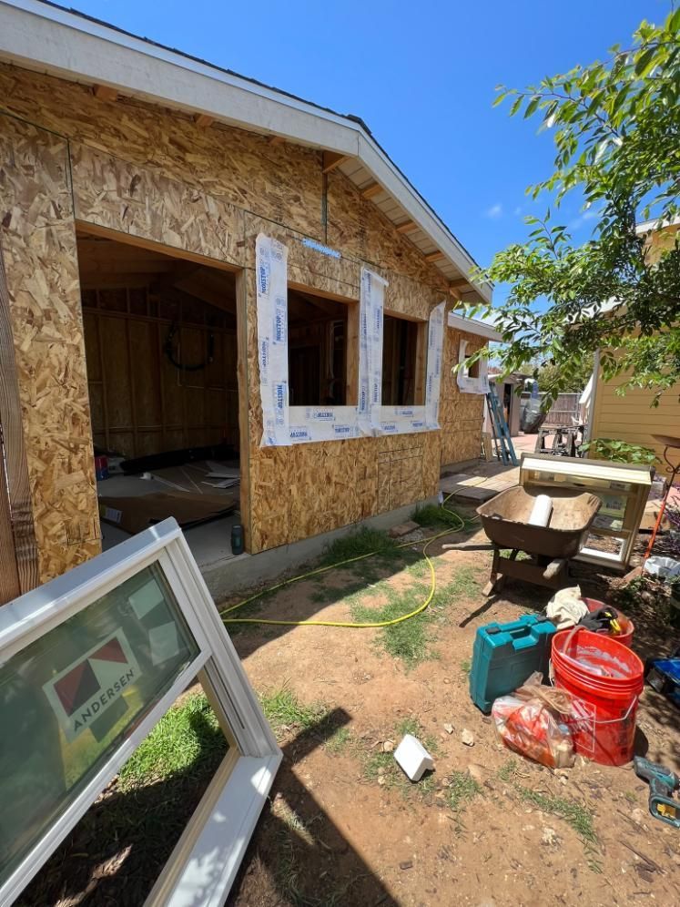 Construction site beside unfinished house with plywood siding, windows, tools, and debris in a sunny yard