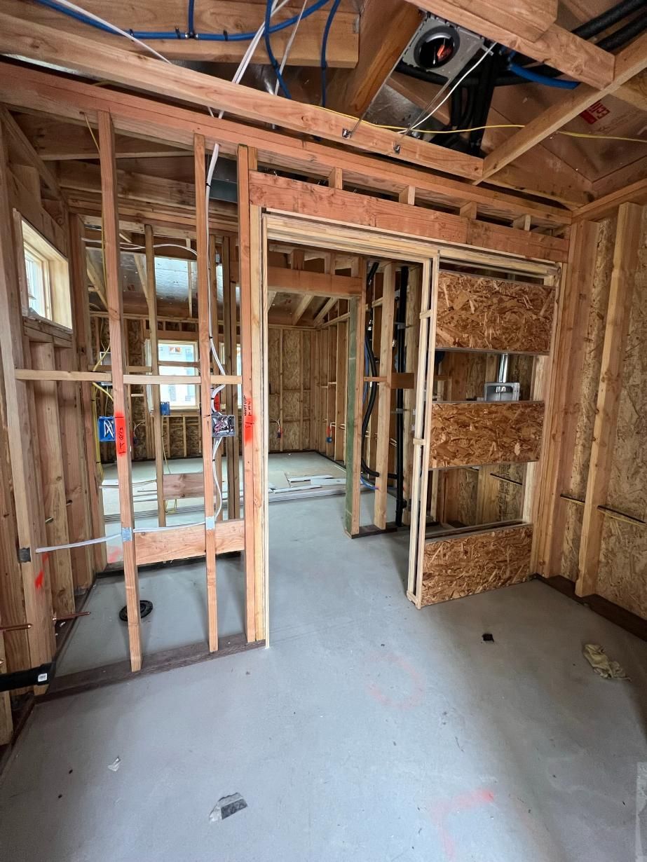 Interior of a house under construction with exposed wooden framing, concrete floor, and a doorway to another room.