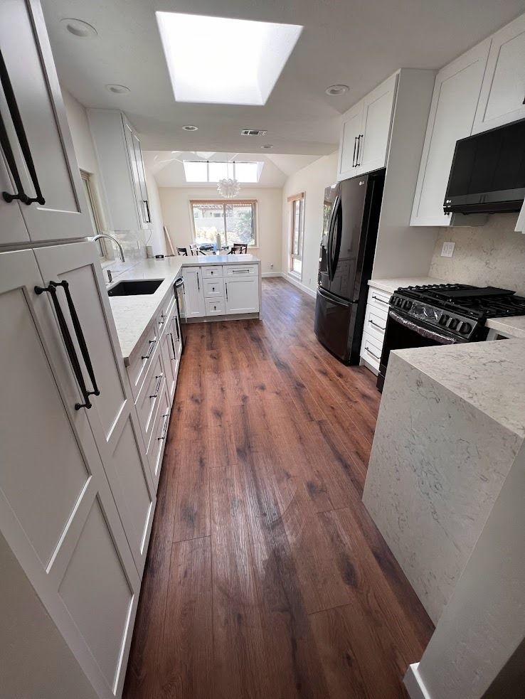 Bright modern galley kitchen with white cabinets, skylight, black appliances, and wood floors