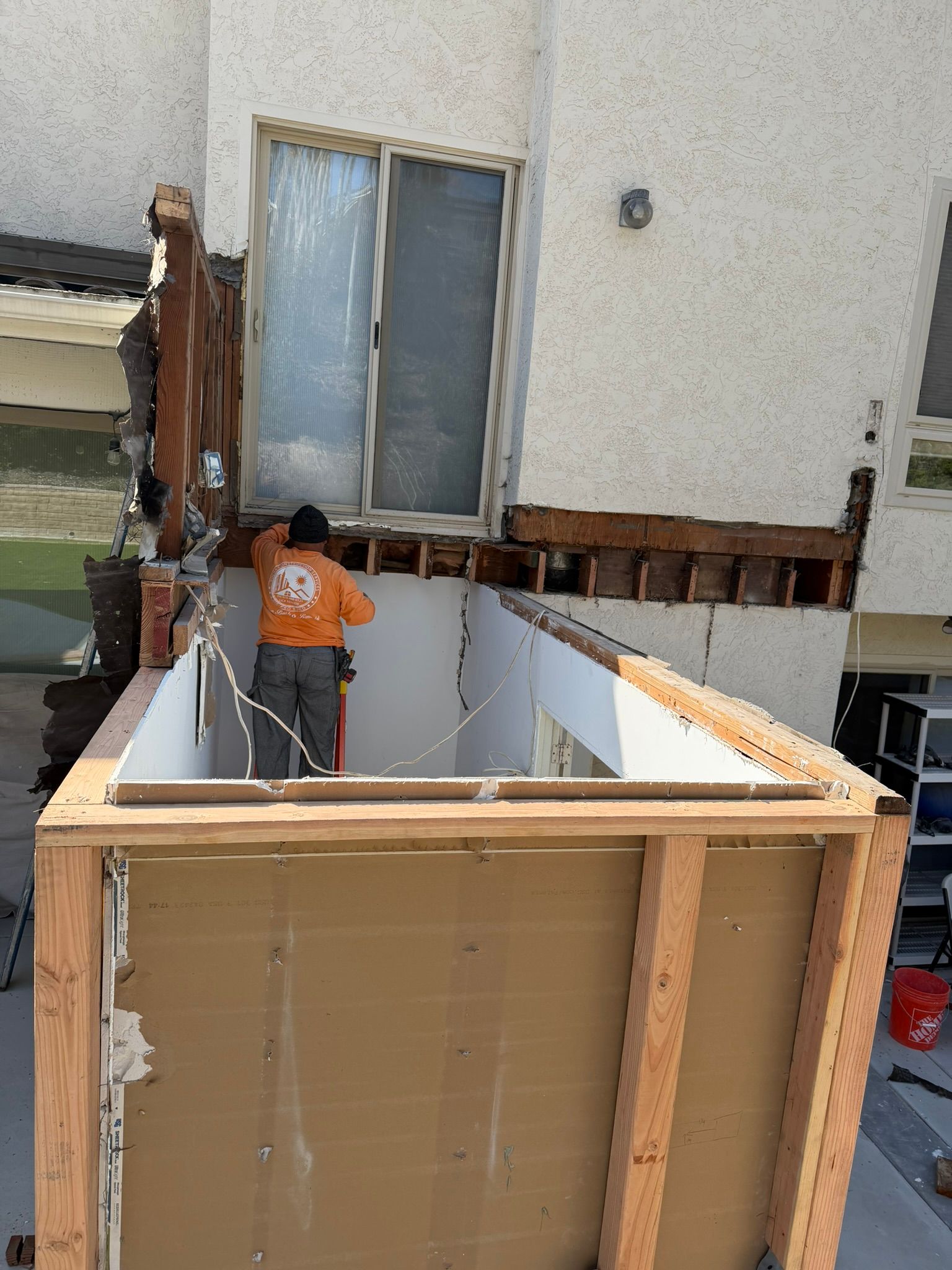 Worker on a small balcony construction site beside a house, with wooden framing and white panel walls.