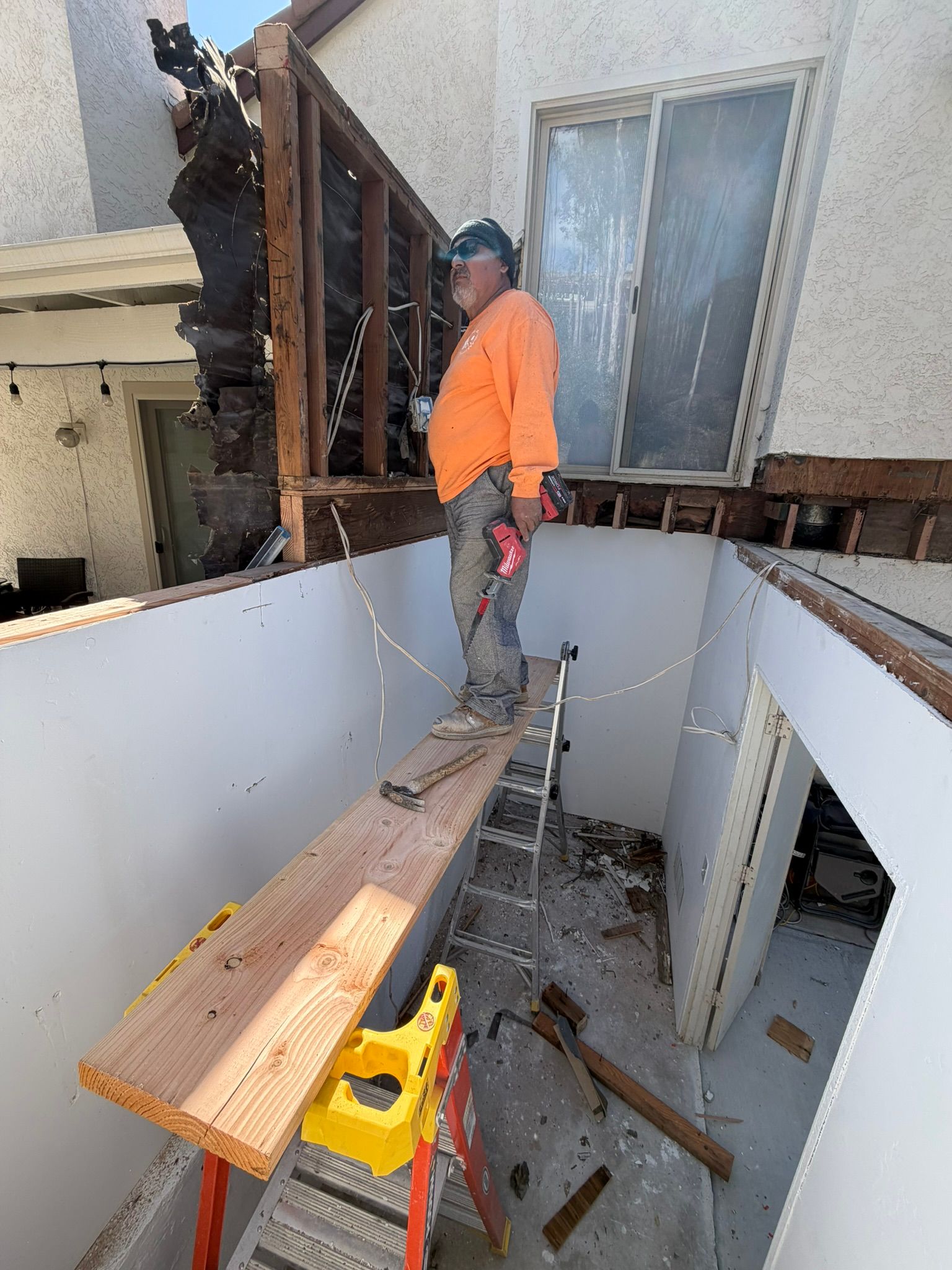 Worker in orange shirt on a narrow plank inside a damaged room with exposed framing and debris