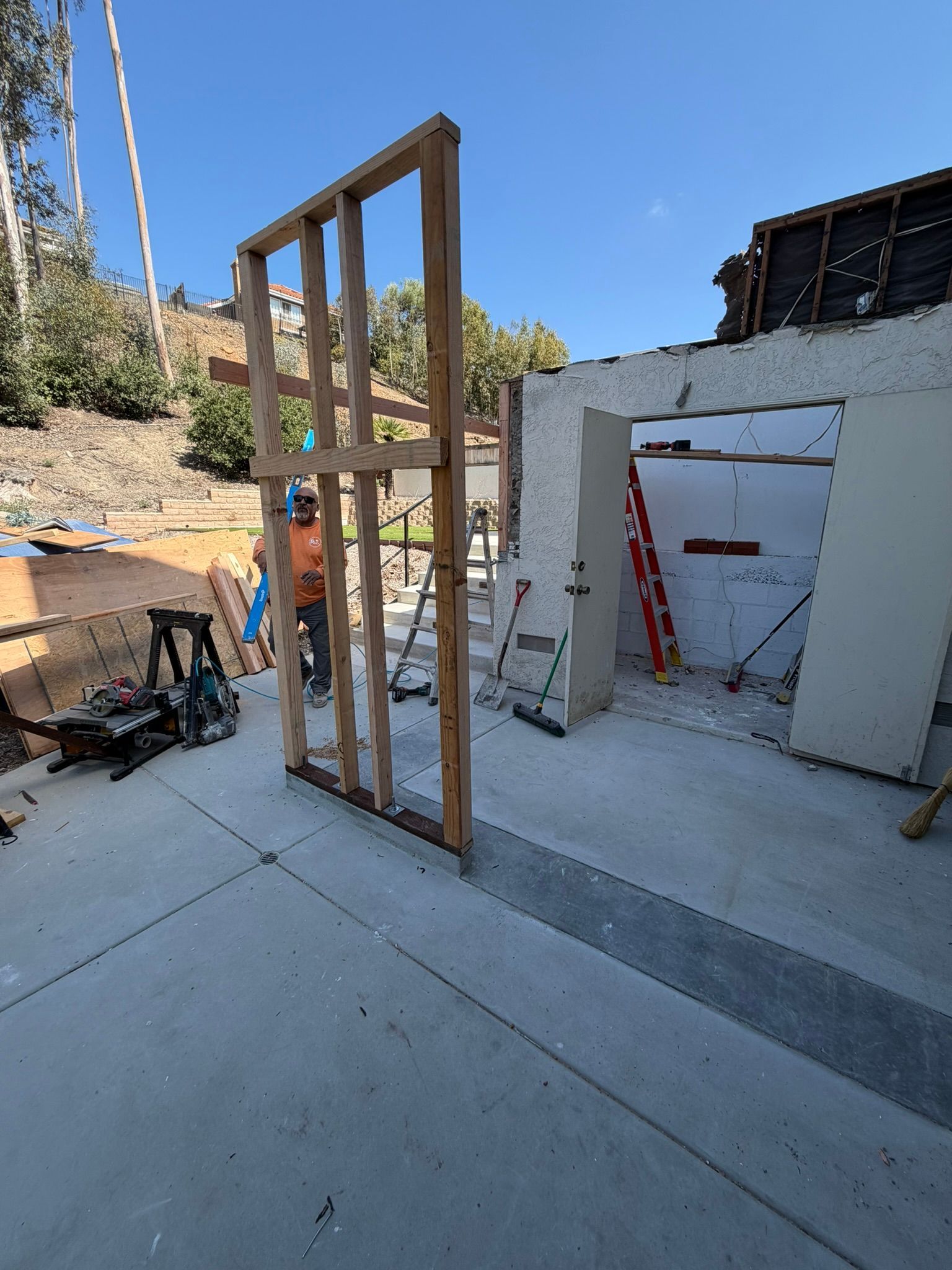 Partially framed wall under construction beside a white stucco structure on a concrete patio, with tools nearby