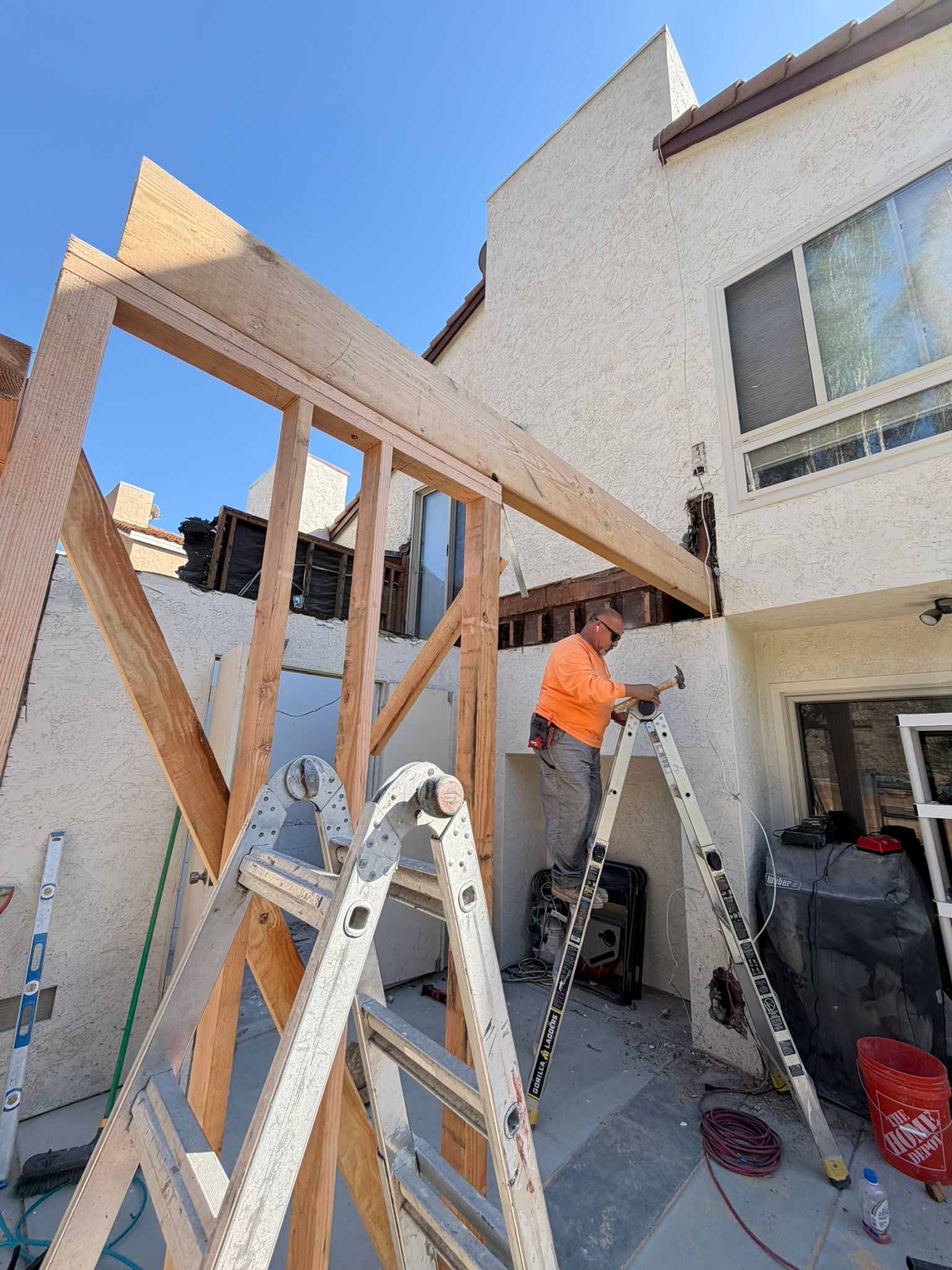 Worker building a wood frame addition beside a white house, with ladders and tools in the yard