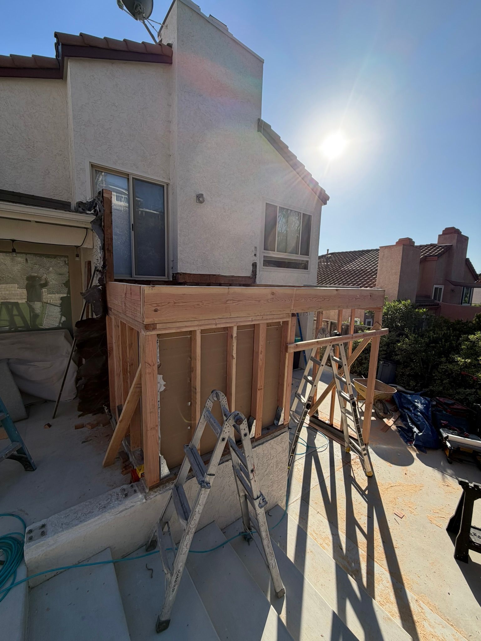 Partially built wooden deck under construction beside a stucco house, with ladders and bright sunlight.