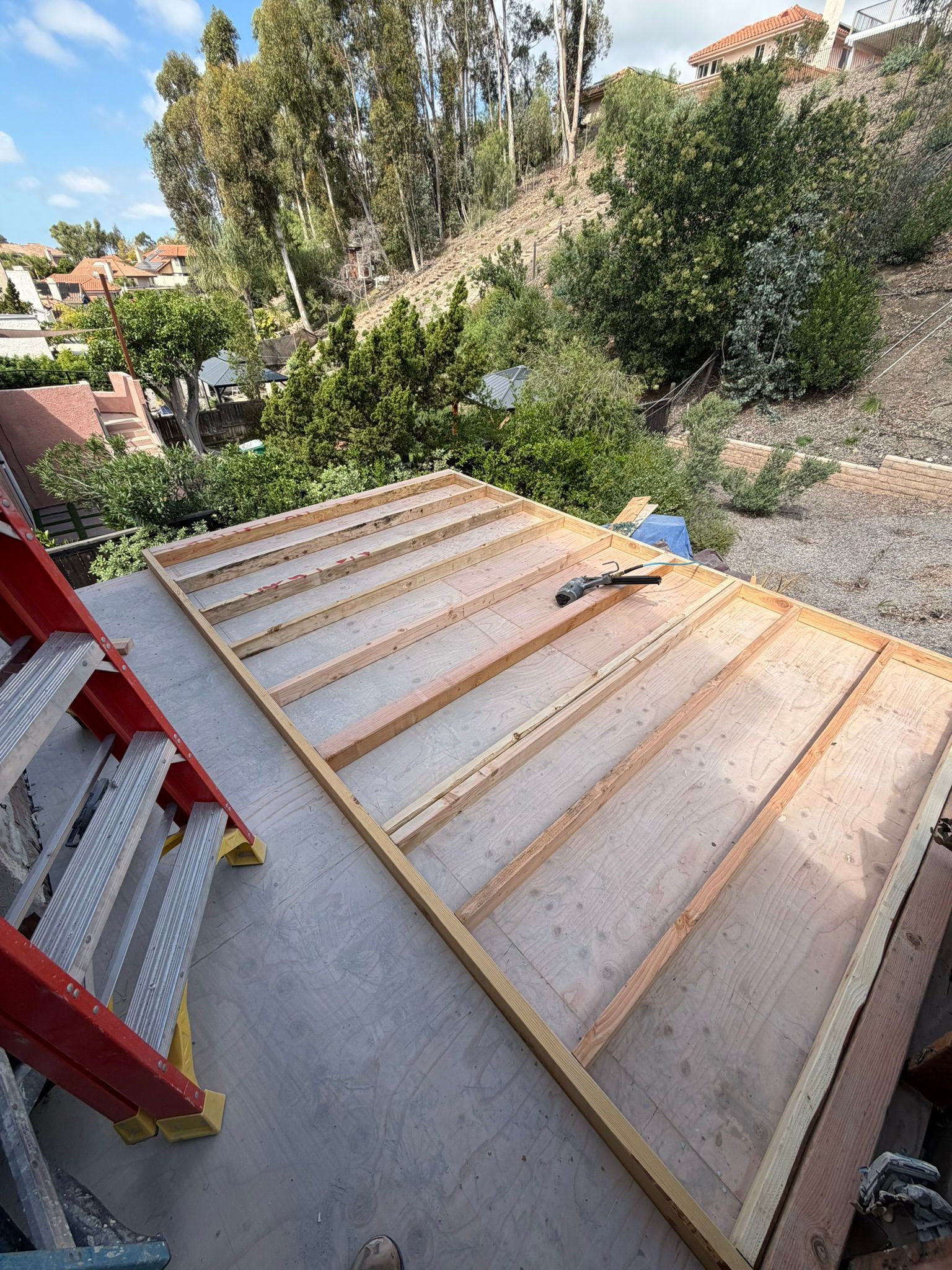 Wooden roof frame laid on a concrete patio beside a red ladder, with hillside greenery beyond.