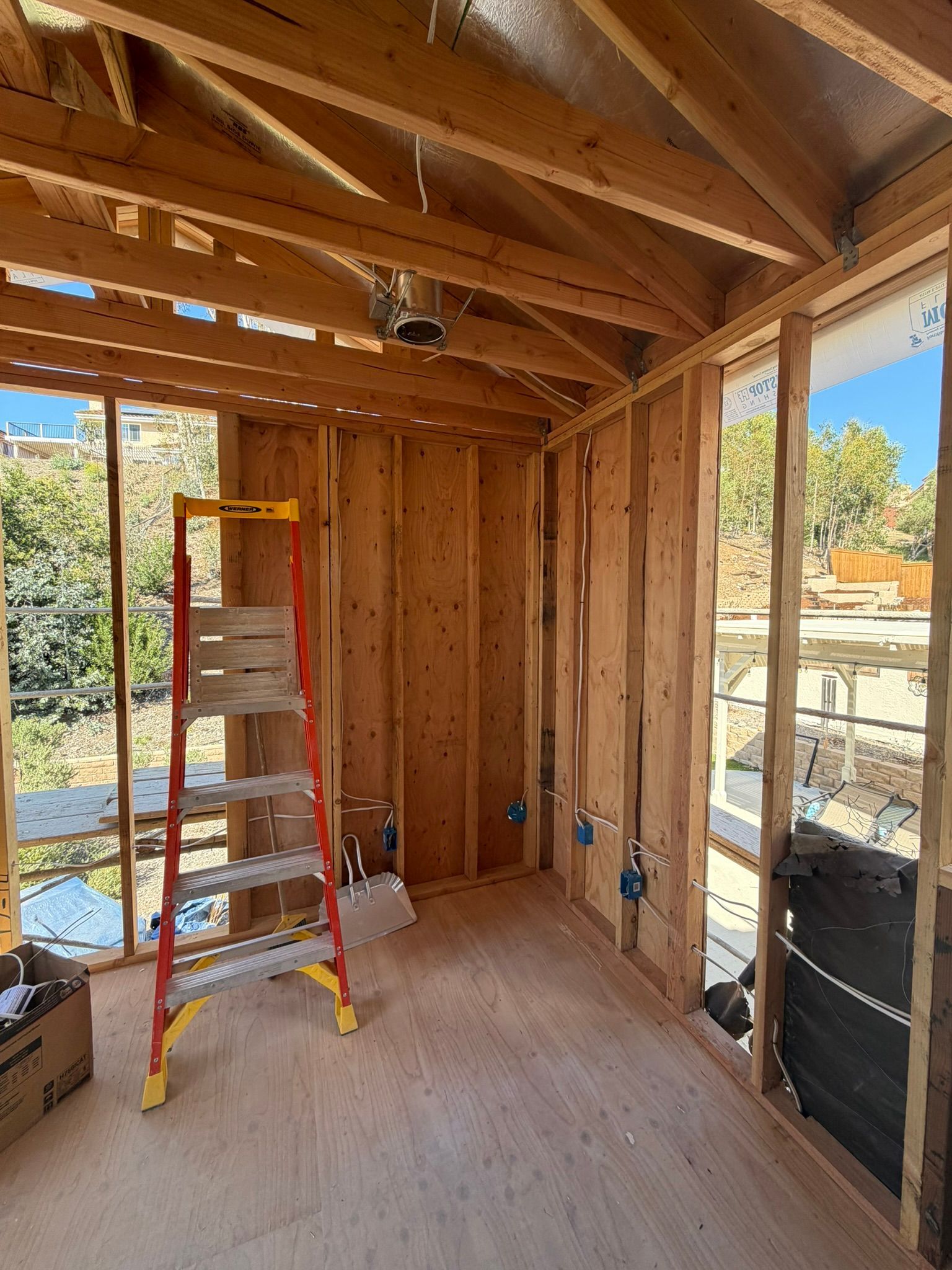 Framed room under construction with exposed wood beams, ladder, plywood floor, and windows overlooking a yard