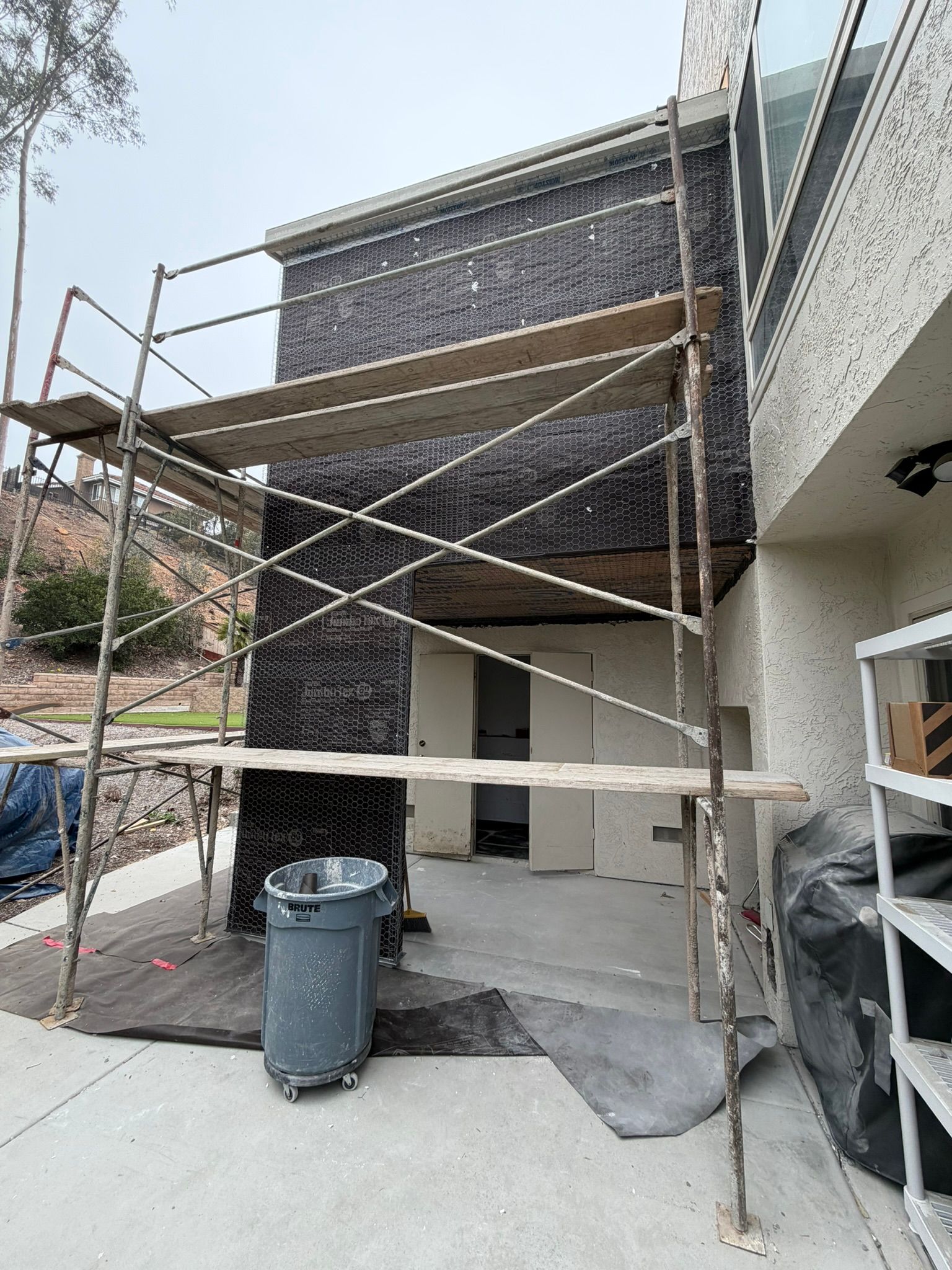 Scaffolded building facade with dark netting, construction materials, and a trash bin in the foreground