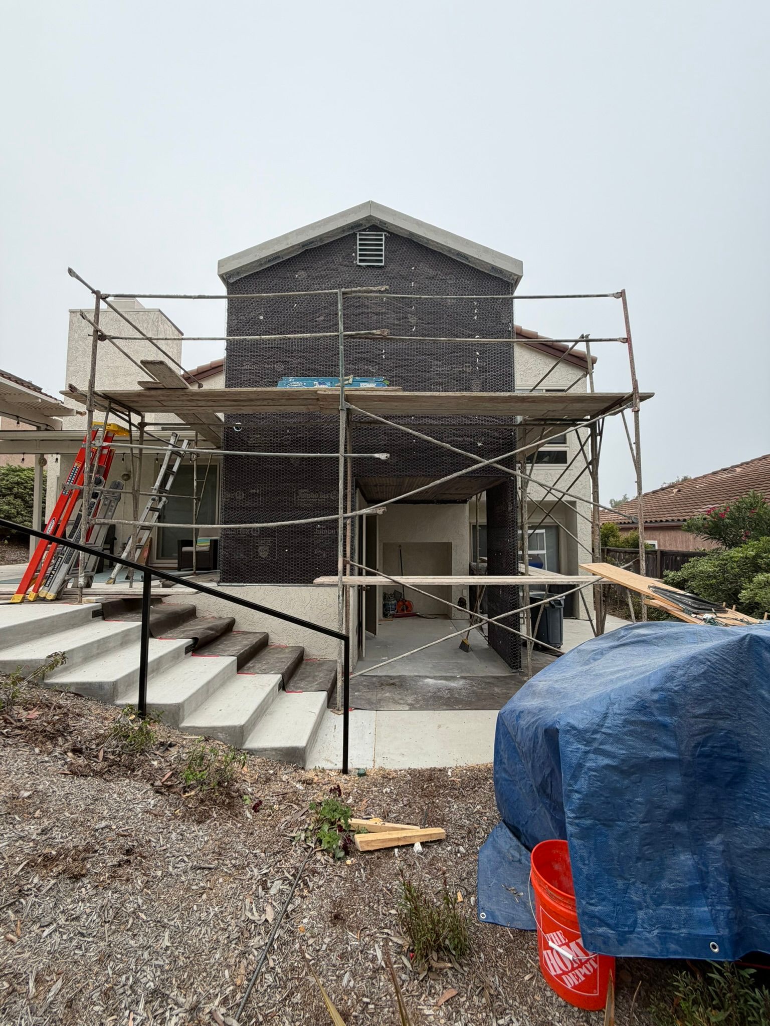 Brick house under construction with scaffolding, steps, and a blue tarp in front.