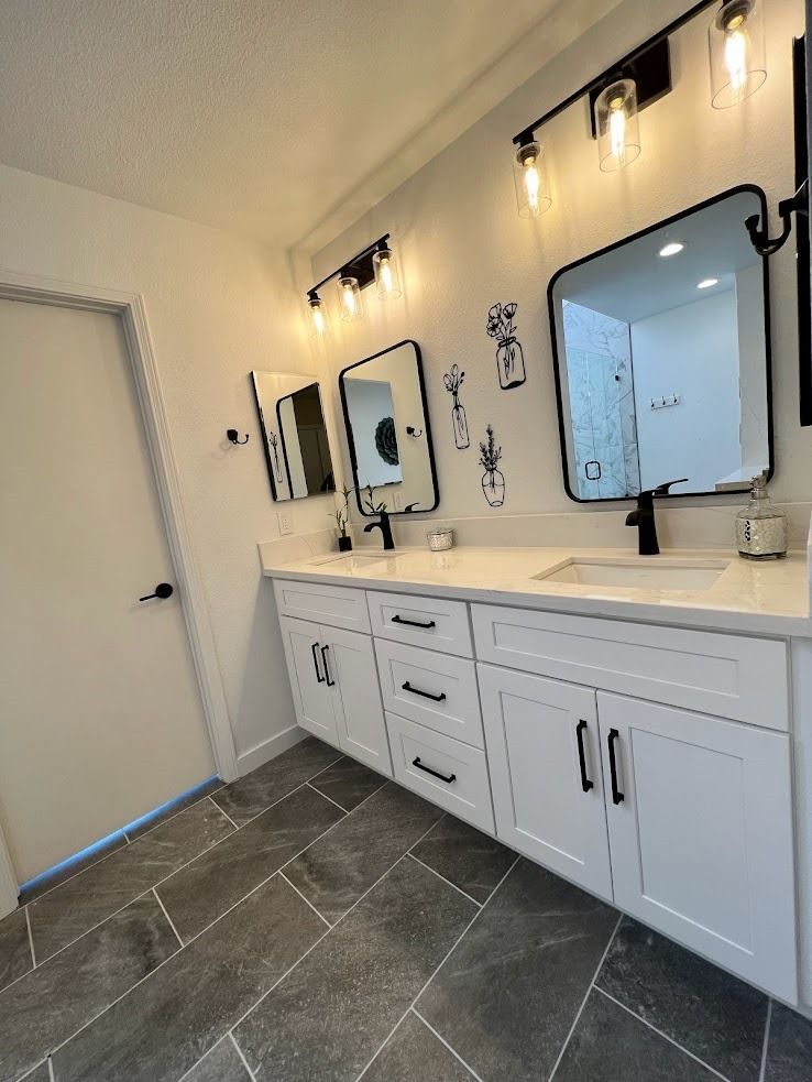 Modern bathroom vanity with white cabinets, black fixtures, mirrors, and wall sconces on a gray tile floor