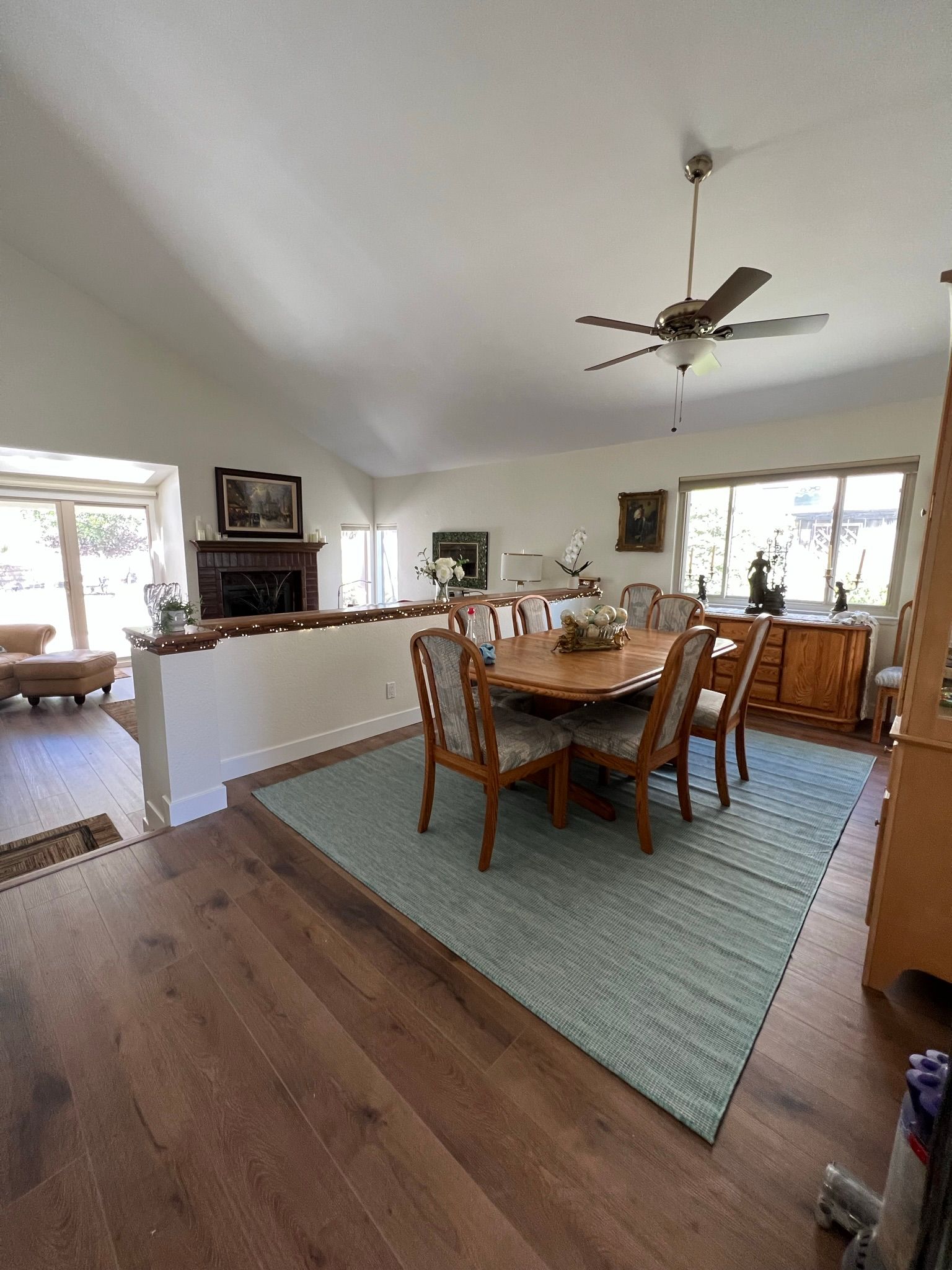 Dining room with wooden table and chairs on a blue rug, open to a bright living area and windows.