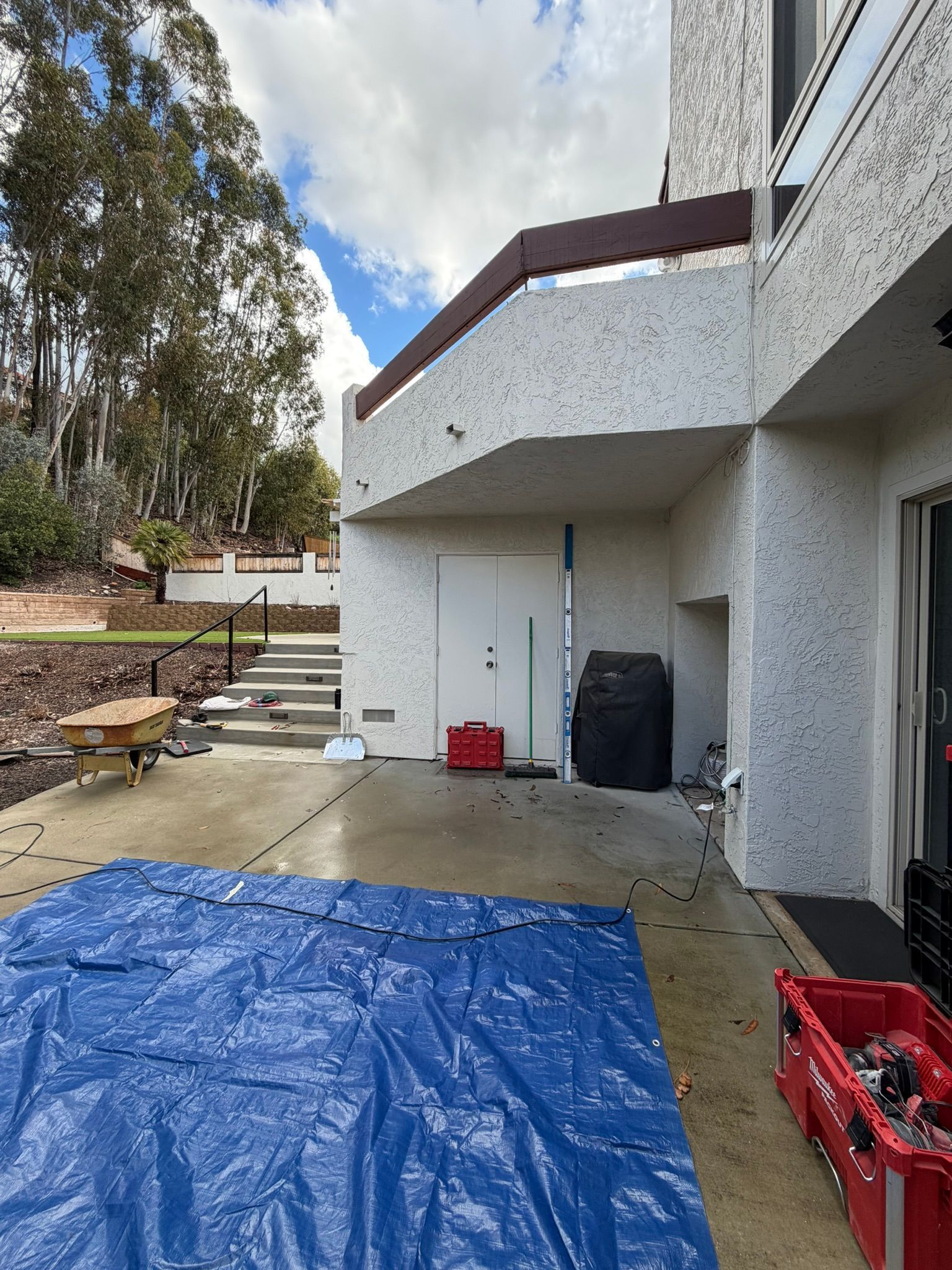 Backyard patio with blue tarp, stairs, red tools, and a white house exterior