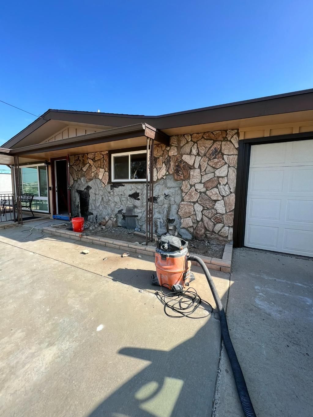 Stone-front house exterior with white garage door, concrete driveway, and patio furniture in bright sunlight