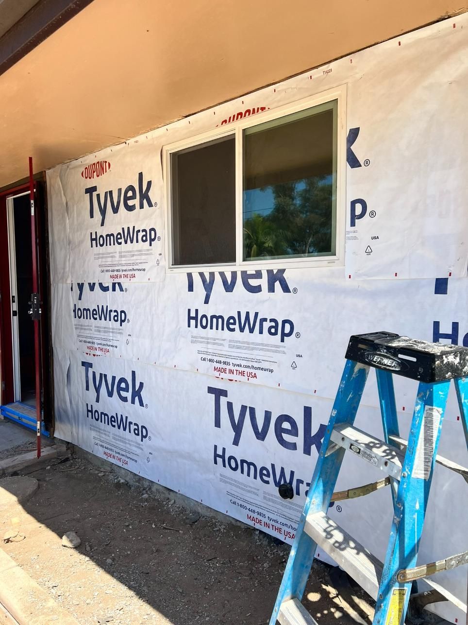 House under renovation with Tyvek wrap, a window opening, and a blue ladder in the foreground