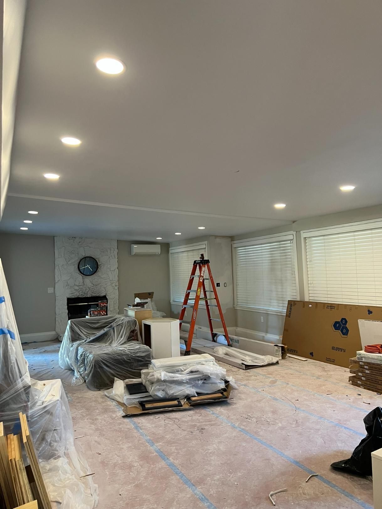 Renovation in progress in a bright living room with ladder, covered furniture, and floor protection