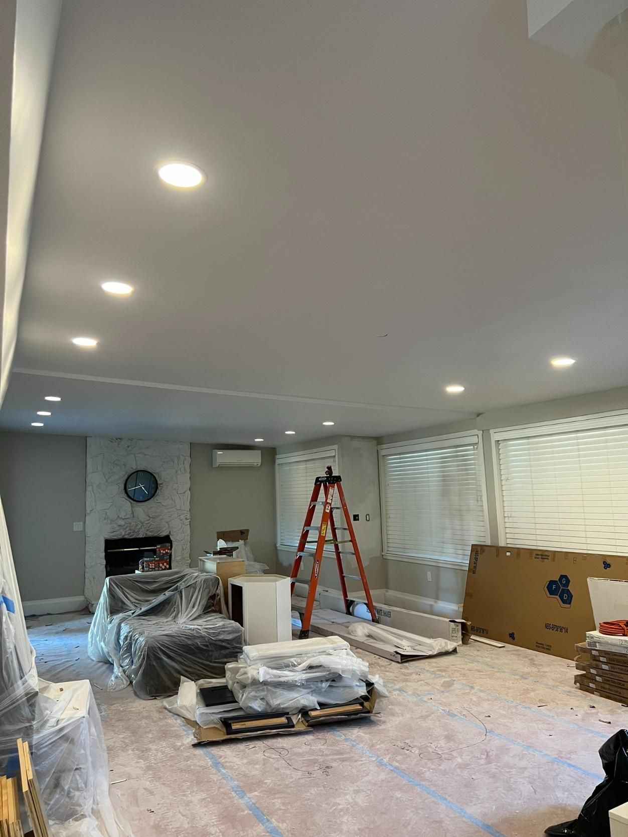 Living room under renovation with plastic-covered furniture, ladder, tools, and recessed ceiling lights.