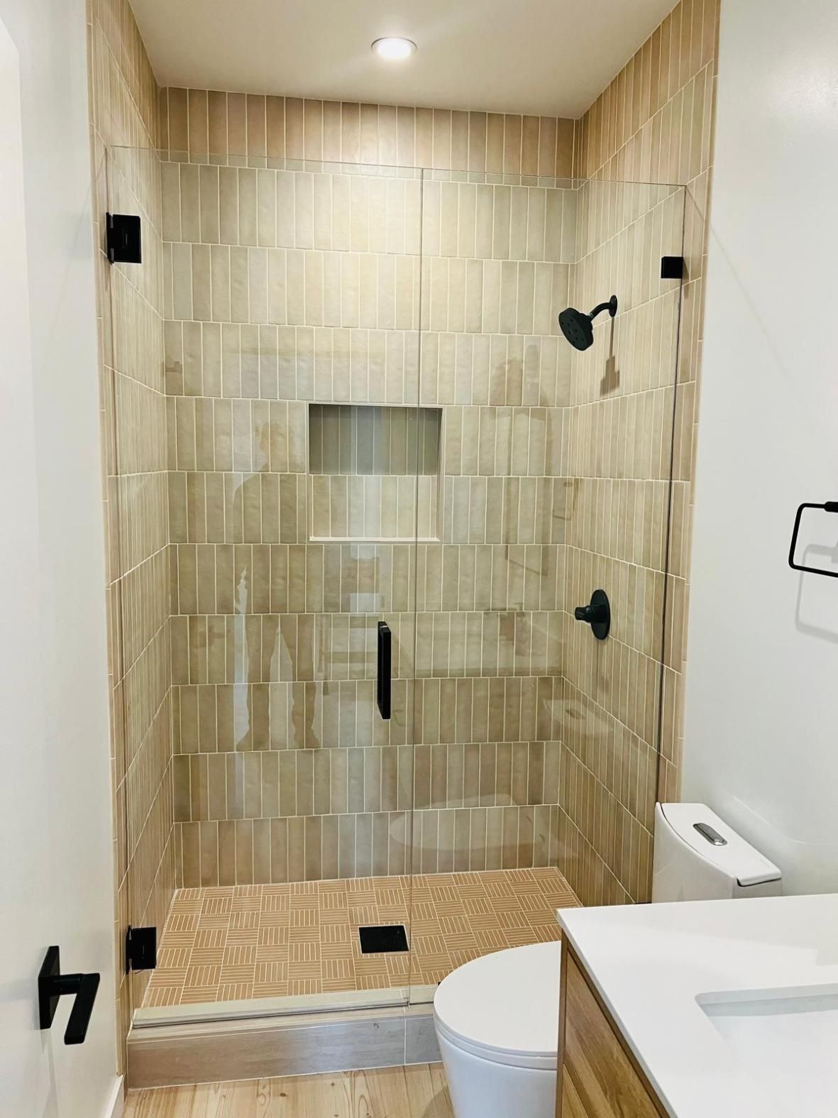 Modern beige-tiled walk-in shower with glass door, bench, built-in niche, and adjacent toilet.