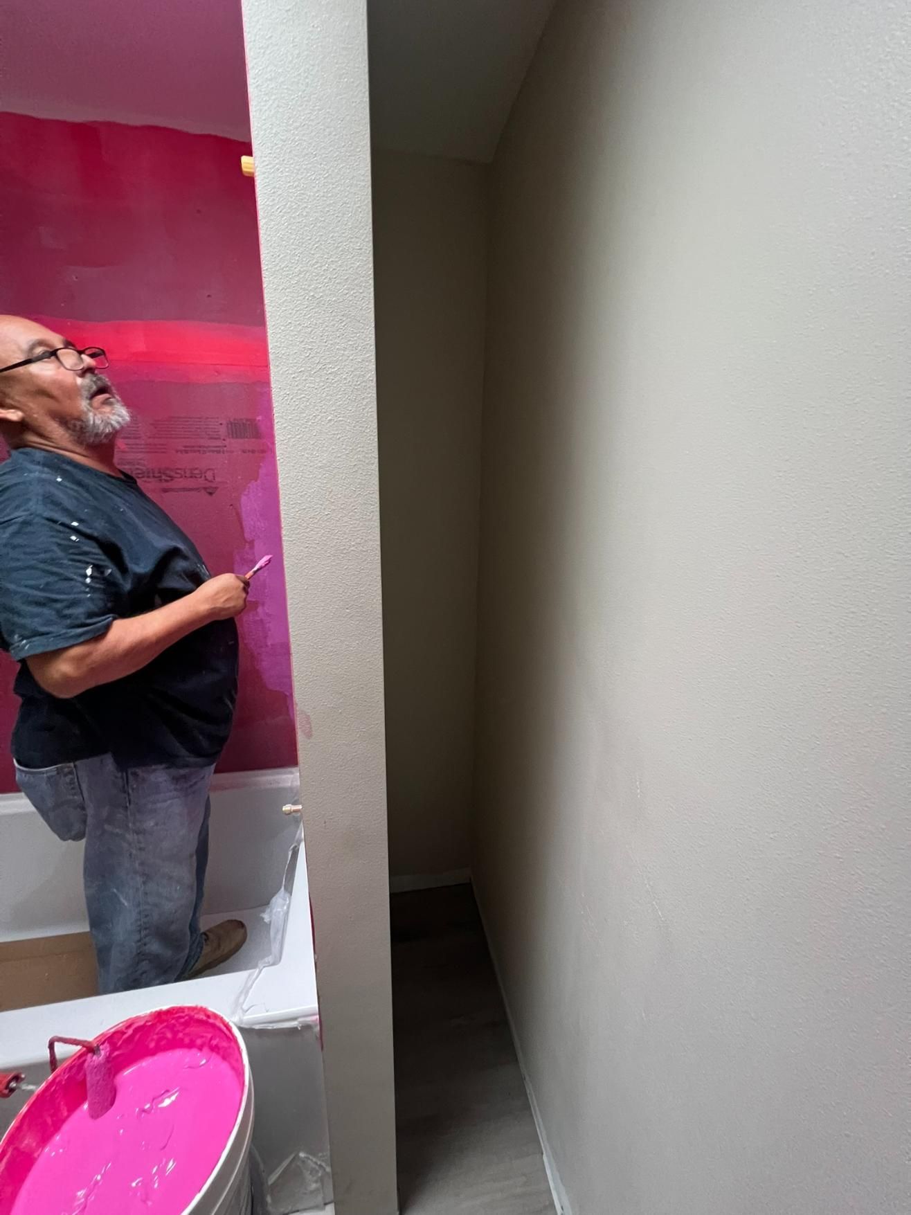 Man in a bathroom stall beside a pink sink, with a bright pink wall panel behind him.