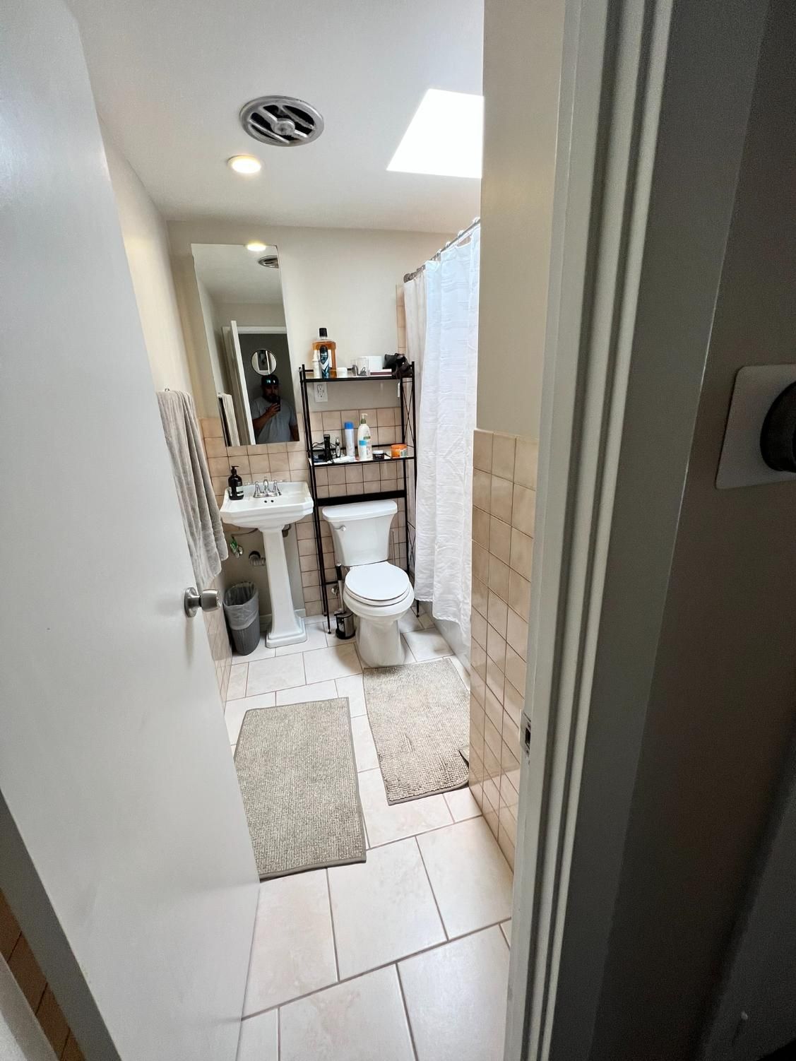 Small bathroom with white tile, toilet, sink, shower curtain, and a shelf of toiletries.