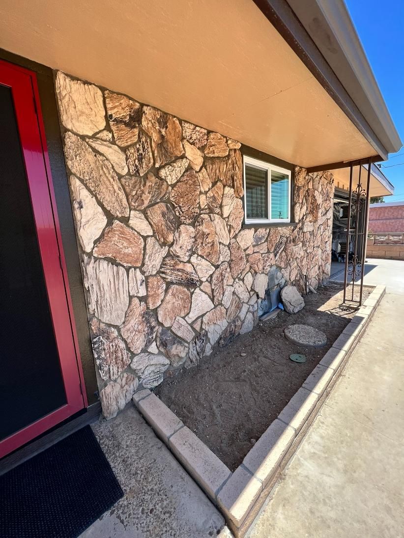Stone exterior wall beside a red door and small window along a narrow walkway in bright sunlight