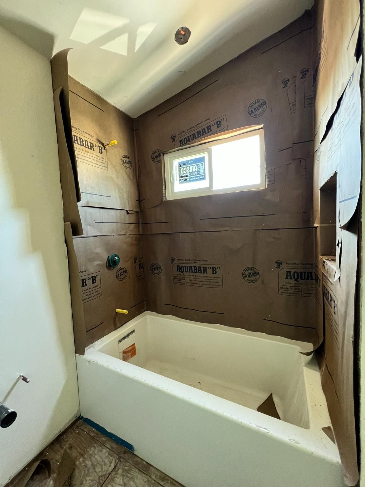 Unfinished bathroom with a white bathtub, partially tiled walls, and a small window letting in daylight