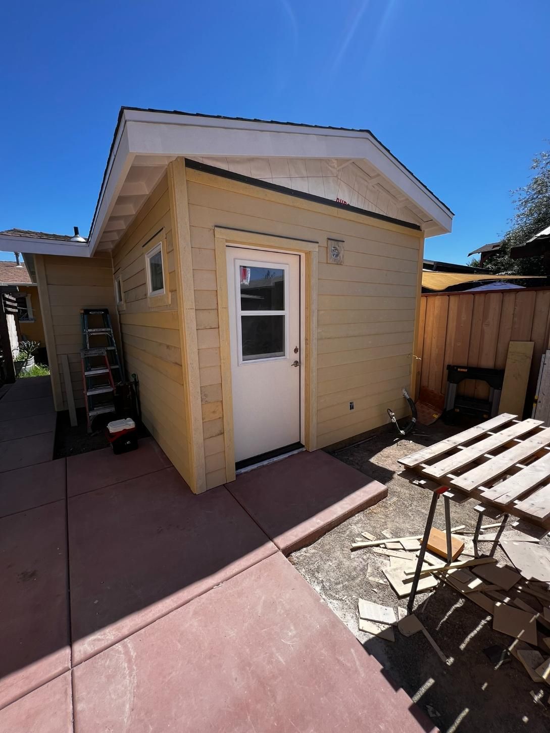 Small yellow backyard shed with a white door beside a patio and wooden fence under a blue sky