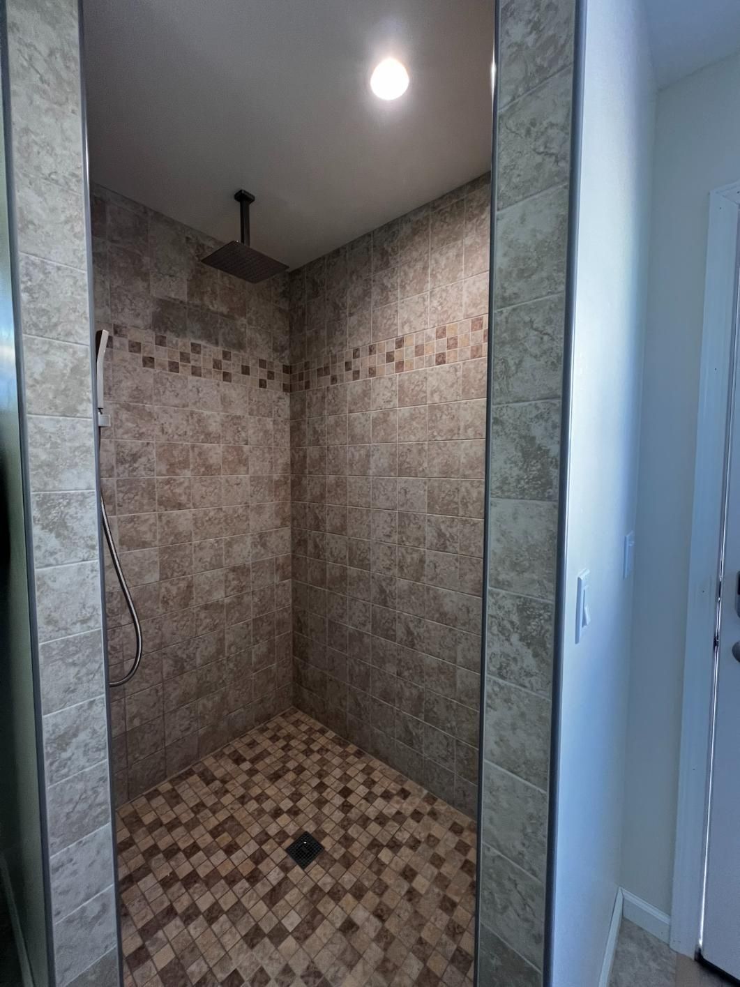 Empty tiled shower stall with beige mosaic walls and floor, lit by a ceiling light.