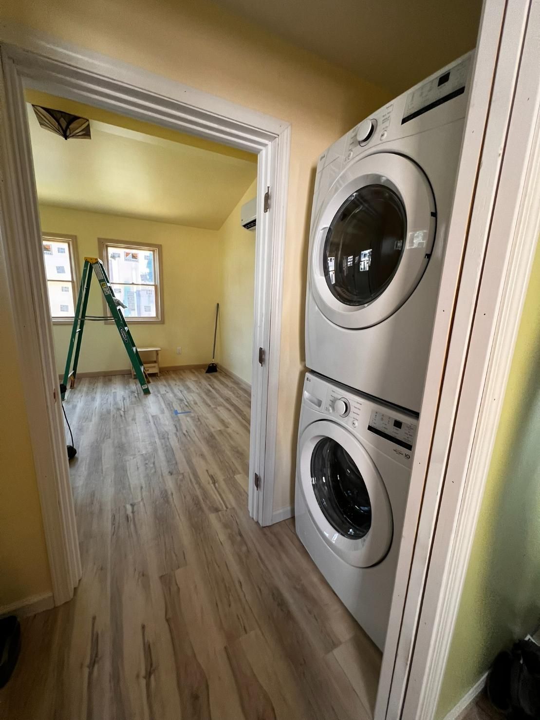 Laundry room with stacked washer and dryer, wood floors, and a ladder in the adjoining room.