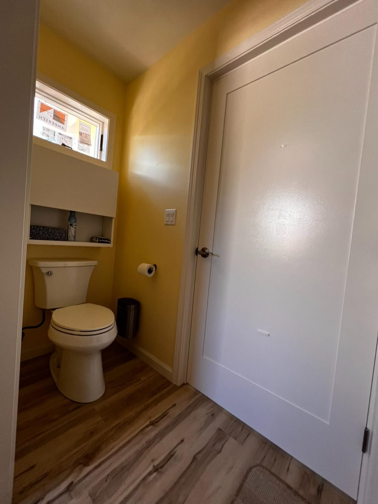 Small yellow bathroom with toilet, sink alcove, and white door on wood-pattern floor.