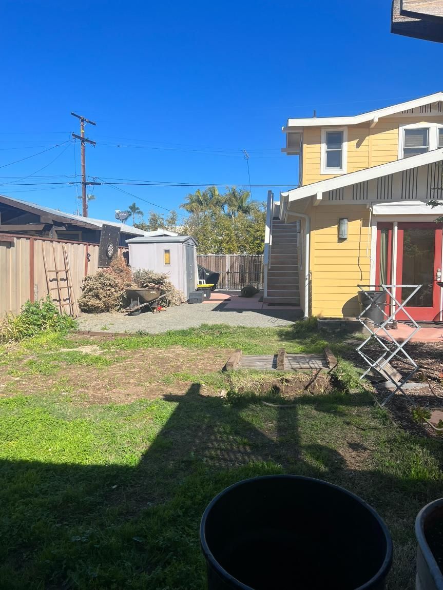 Sunny backyard with yellow house, shed, grassy yard, and patio chairs under a blue sky