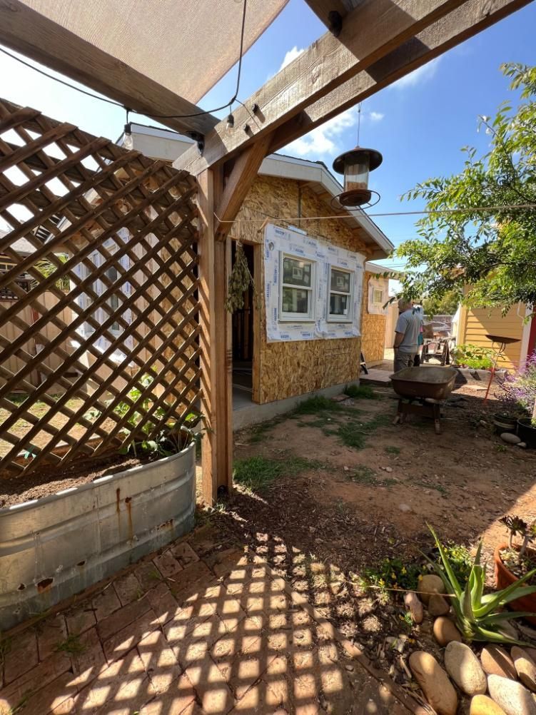Sunlit backyard with a lattice trellis, stone house wall, garden path, and potted plants under blue sky