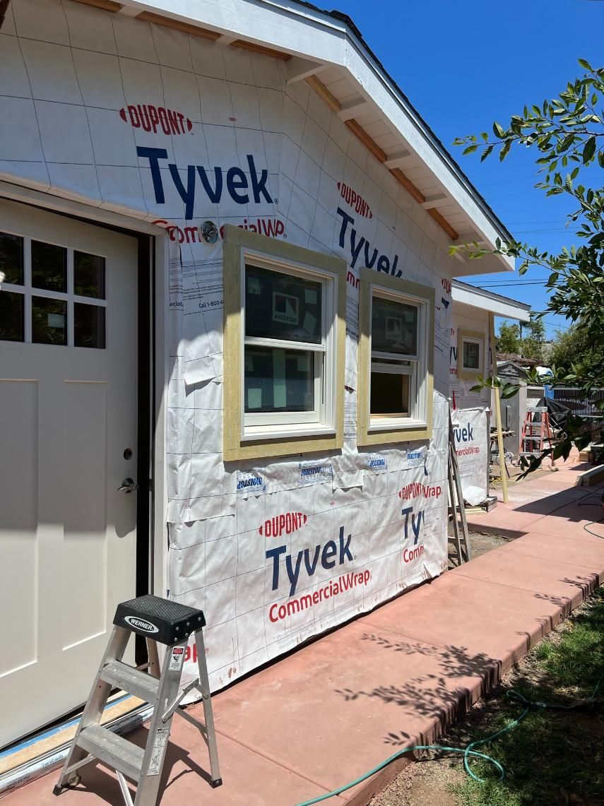 House exterior under construction wrapped in Tyvek, with a ladder by the door and window framing in progress