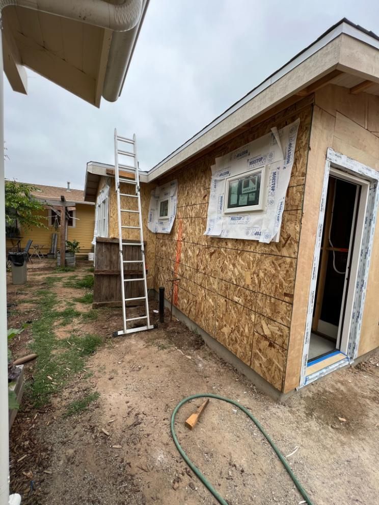 Partially built house with OSB siding, open doorway, and ladder in a narrow dirt yard