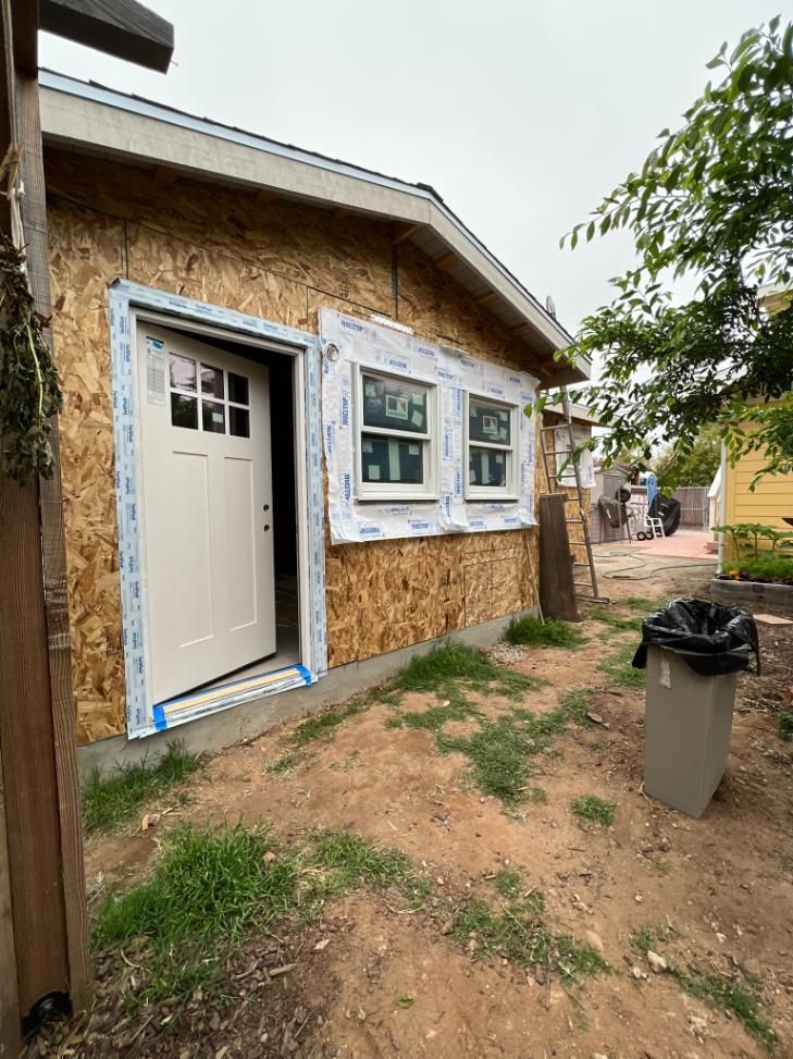 Small stucco shed with open white door and windows beside a dirt yard and trash bin